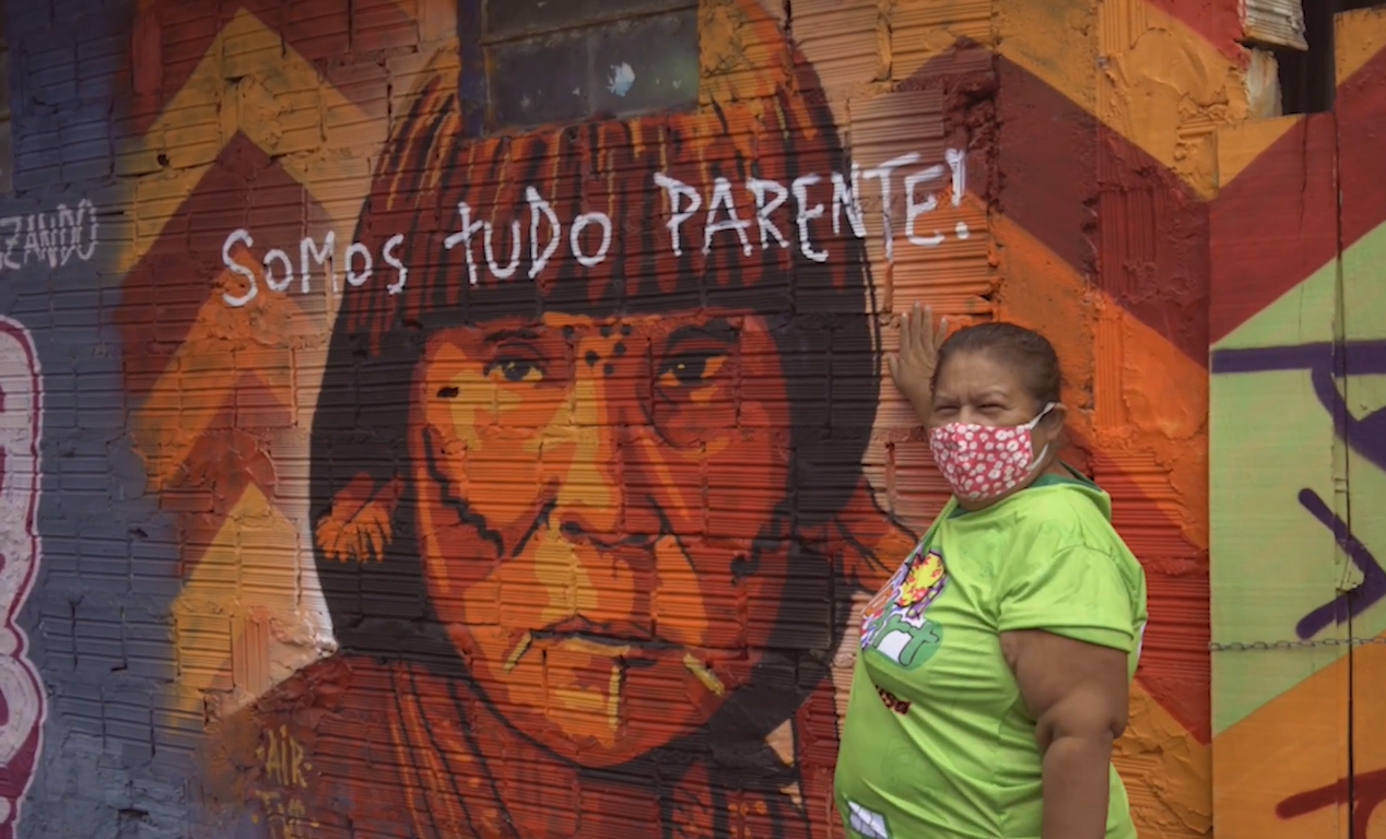 Mulher próxima a uma parede pintada com a foto de um indígena e com a frase "somos tudo parente!".