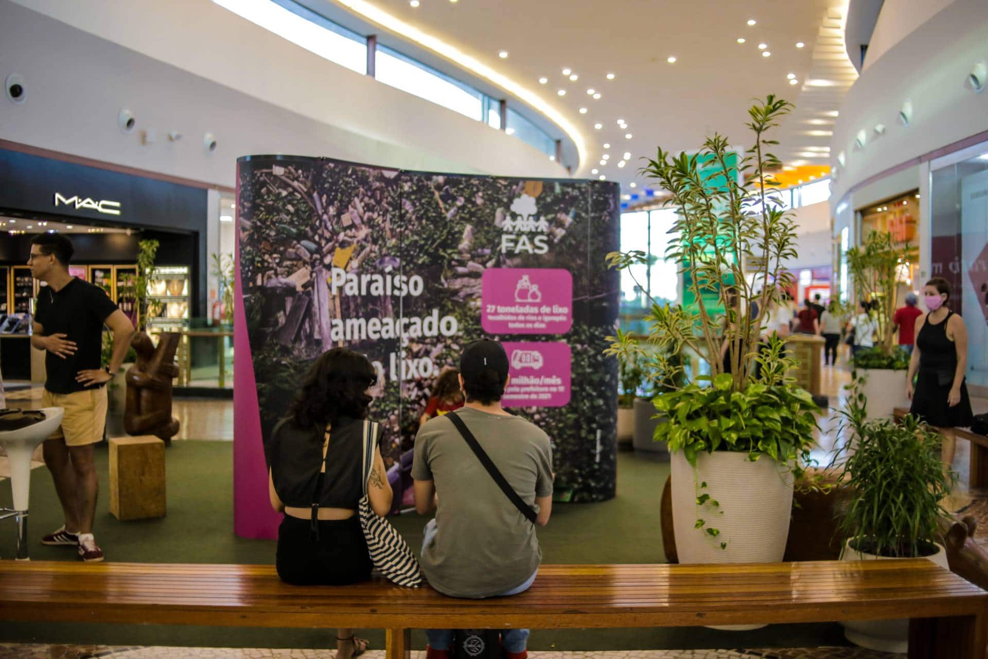 Pessoas sentadas em banco no shopping, vendo exposição realizada pela Fundação Amazônia Sustentável (FAS).