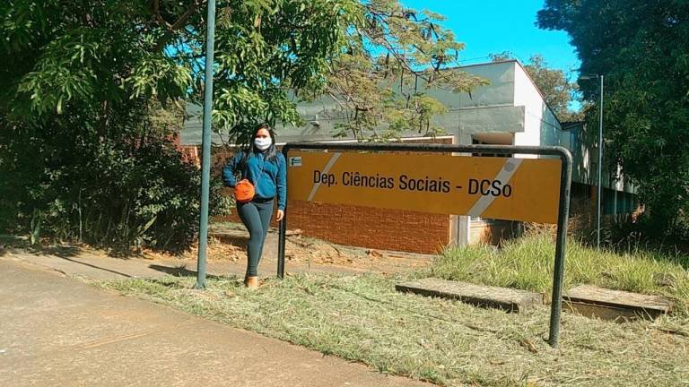 Mulher posando para foto ao lado de uma placa dentro da Unicamp-UFSCar.