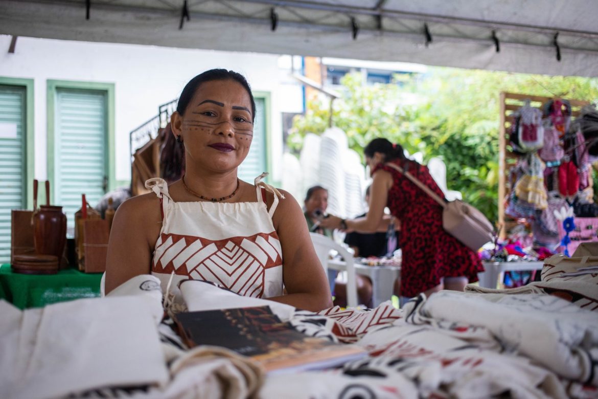 Mulher indígena sentada próximo ao seu stand, participando da Feira da FAS, realizada pela Fundação Amazônia Sustentável (FAS).