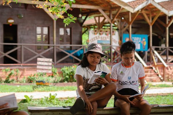 Crianças sentadas em banco, lendo livro em comunidade no interior do Amazonas.